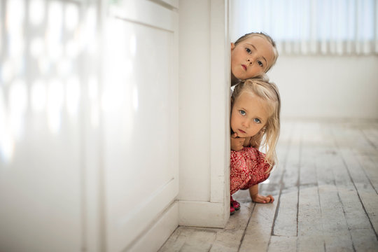 Two young blond sisters peek around the edge of a doorframe.
