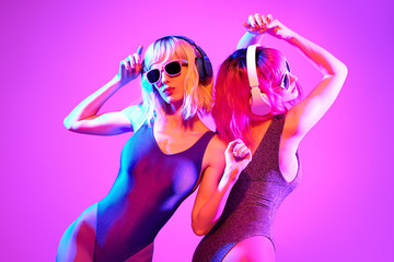 Two shapely fashionable DJ girl dance enjoy music in colorful neon uv purple blue light. Rave house music night club vibes. High Fashion. Young model woman friends relax, neon makeup.
