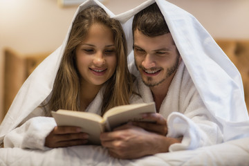 Beautiful woman and handsome man lying on bed covered with white sheet. Sweet couple holding book in hands and reading. Woman and man in love resting and relaxing in suite.