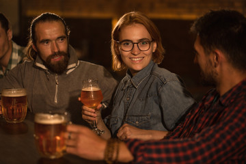 Two men and cheerful woman with ginger hair sitting in beer pub and resting. Beautiful woman wearing in spectacles looking at camera, smiling and holding glass of beer.