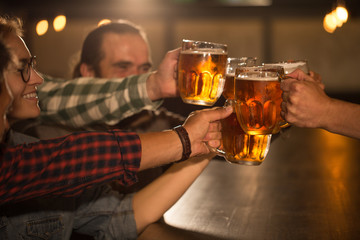 Close up of cheerful company clinking their glasses and pints of beer. Friends hanging out and resting in beer pub or bar. Hands holding glasses full of delicious lager.