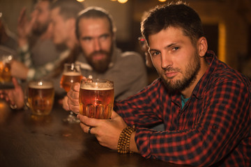 Client of beer pub sitting and resting. Handsome bearded man in checked shirt looking at camera and holding glass of tasty beer. People in pub communicating and socializing.