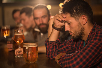 Sad and depressed facial expression of bearded man looking down. Male client sitting in beer pub and holding hand on head. People resting in bar and communicating on background.