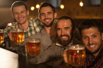 Four friends looking at camera and posing with glasses of tasty beer they holding in hands. Handsome and cheerful men smiling. Company resting and having meeting in beer pub.