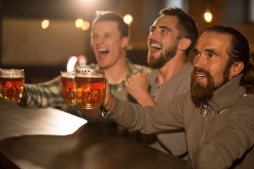 Three male friends looking up, shouting and smiling. Positive and happy men watching football and enjoying weekend. Clients of pub holding glasses of beer and hanging out together.