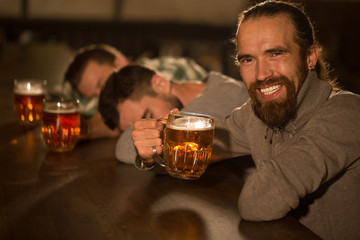 Side view of positive and happy bearded; handsome man holding glass of beer and drinking. Man enjoying and tasting delicious beer looking and smiling at camera. People resting in pub.