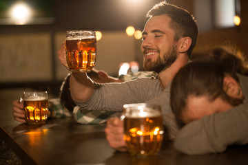 Funny men holding glasses of beer and sitting in pub. Young bearded man smiling and looking at beer glass. Two men lying on table and sleeping. Friends hanging out in bar.