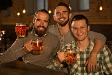 Front view of three friends posing together with glasses of beer, they holding in hands. Handsome and cheerful men smiling and looking at camera. Company hanging out together in beer pub.