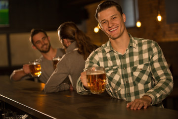 Handsome man smiling, looking at camera and holding glass of beer in hand. Young cheerful man in checked shirt posing. Male clients sitting and hanging out in beer pub.