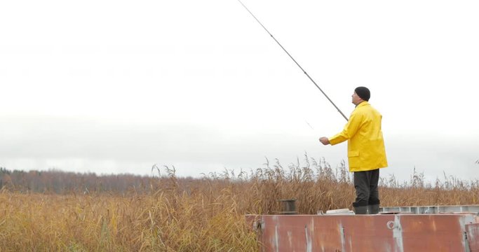 Handsome plus size young man in yellow fisherman coat jacket. A man is fishing from a metal pier with fishing rod at beautiful big lake. Rural getaway concept.