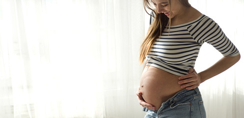 Happy Attractive pregnant woman standing near the window and holding her belly