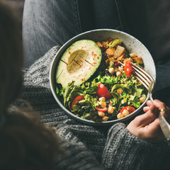Healthy vegetarian dinner. Woman in jeans and woolen sweater holding bowl with fresh salad, avocado...