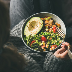 Healthy vegetarian dinner. Woman in jeans and woolen sweater holding bowl with fresh salad, avocado half, grains, beans, roasted vegetables, top view, square crop. Superfood, clean eating food concept