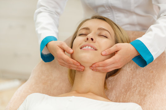 Beautiful female lying on couch and enjoying massage after peeling in cosmetology salon. Skilled doctor in uniform doing relaxing procedure zo young client for removing wrinkles and skin comfort.