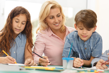 Fototapeta premium blonde woman sitting at table between two drawing pupils and smiling.