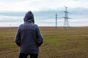 Conceptual anonymous unidentified person in hood is standing on field in front of electric pylon and looking