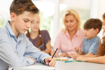 Fototapeta premium Boy with serious facial expression, sitting at desk and drawing pencil on paper.