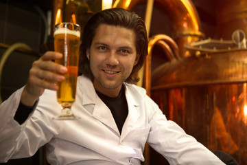 Handsome brewer showing glass of tasty beer and looking at camera. Specialist of brew industry smiling and posing. Man working in brewery with brew kettles and other machinery.