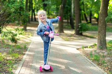 Cute little blond kid girl riding scooter in city park. Preschooler girl in jeans overall having fun during outdoor sport activities. Summer leisure and recreation time