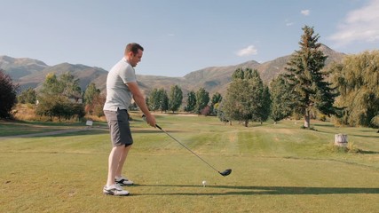 Middle Aged Man Tees Off On Golf Course. The video shows a middle aged man hitting a golf ball. Trees and mountains in the background.