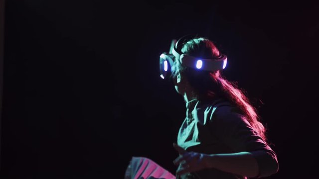 Young woman wearing VR headset making gesture in abstract dark space with neon lights
