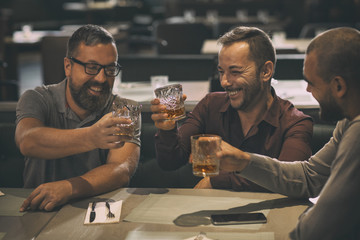Cheerful smiling men holding glasses with alcohol beverages and laughing. Three friends spending time together and having fun in bar. Men drinking scotch, whiskey or brandy.