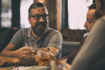 Bearded man in spectacles with glass in hand looking at his friends. Men sitting in bar together, communicating and talking. Clients of bar holding crystal glasses with alcohol.