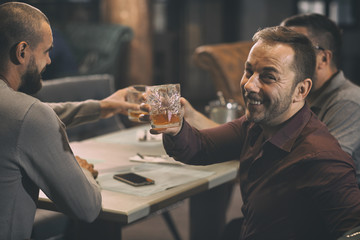 Cheerful man in shirt looking through shoulder at camera and smiling. Happy man holding and showing glass with whiskey or scotch. Men sitting in bar and spending time together.