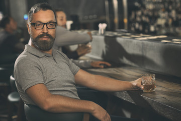 Fototapeta premium Bearded brutal man sitting at counter in bar with loft interior. Handsome man wearing in spectacles and grey t shirt looking at camera and posing. Client of pub holding glass of whiskey in hand.