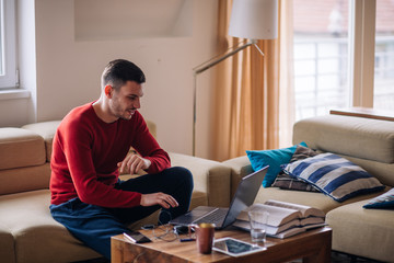 Tired student typing on his laptop and reading a book while sitting on a couch.