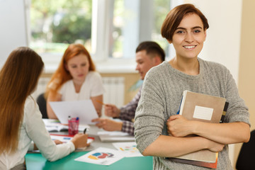 Portrait of young female student keeping book and notes, looking at camera, smiling and posing during lesson on courses. Students studying and talking at background. Interesting learning concept.