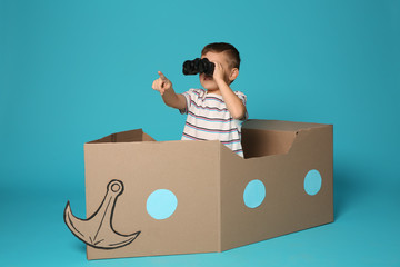 Cute little boy playing with binoculars and cardboard boat on color background