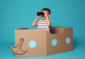 Cute little boy playing with binoculars and cardboard boat on color background