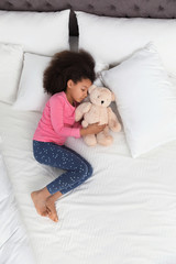 Cute little African-American girl with teddy bear sleeping in bed, top view