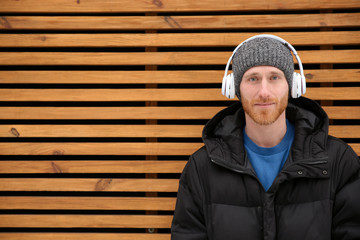 Young man listening to music with headphones against wooden wall. Space for text