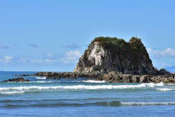 Fototapeta na wymiar Rocky islet surrounded by flat reef bathing in oceanic surf near mouth of Mangawhai Harbour.