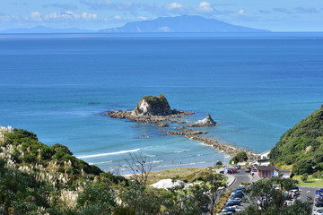 Rocky islet with surrounding reef close to shore with island and distant landmass in far background.