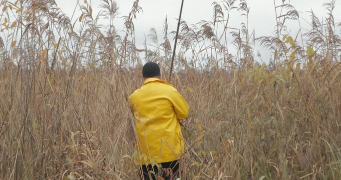 Handsome plus size young man in yellow fisherman coat jacket. Man with fishing rod walk to the lake in tall thickets of dry grass and reeds. Rural getaway concept.