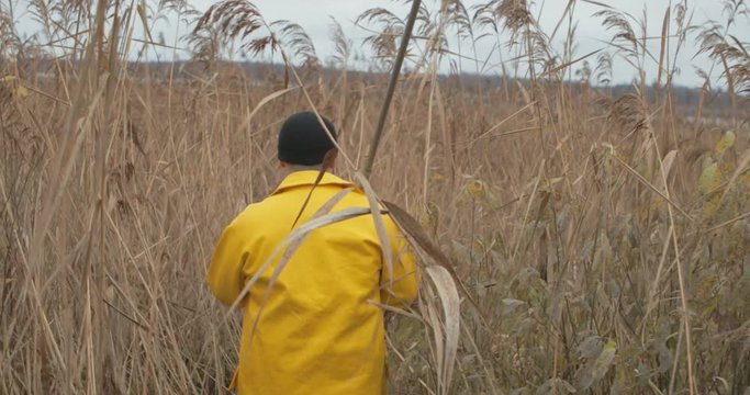 Handsome plus size young man in yellow fisherman coat jacket. Man with fishing rod walk to the lake in tall thickets of dry grass and reeds. Rural getaway concept.