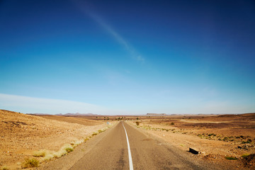 Fototapeta na wymiar A long straight endless road through the desert on a sunny day with blue sky without the clouds in Morocco, Africa