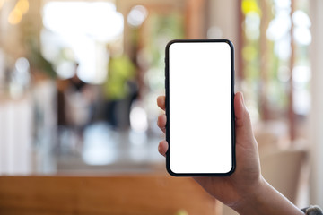Mockup image of a hand holding and showing black mobile phone with blank white screen in modern cafe
