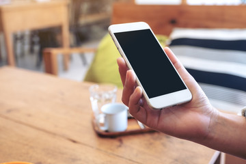 Mockup image of a hand holding white mobile phone with blank screen with coffee cup on wooden table in cafe