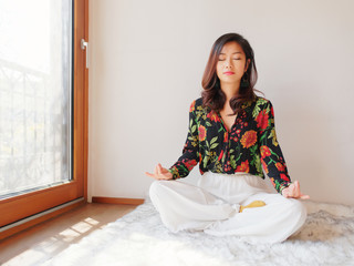 A pretty Chinese young woman practicing yoga at home, sitting on floor with furry cushion near sunny glass door, exercise, Lotus pose, working out, Feeling peace and wellness concept.