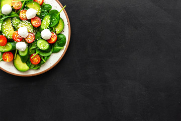 Fresh salad with mozzarella, spinach, cherry tomatoes, cucumber on plate on black background top view space for text