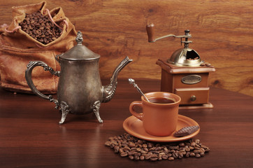 A cup of coffee, coffee grinder, coffee pot, a bag of coffee beans on a wooden table