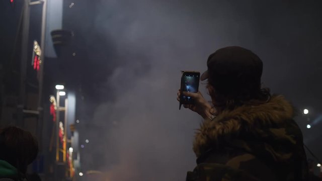 Man filming fireworks of city square. Back view of amn in cap holding smartphone and taking view of bright holiday fireworks on city square in night time.