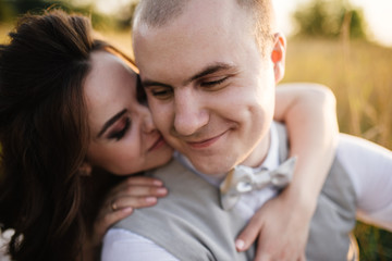 Wedding near the river in field at sunset with brown horse. Bride in light airy dress in color of dusty rose. Beige dress with sparkles. Light suit with bow tie. bride and groom embrace and kiss.