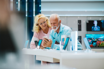 Mature couple leaning on stand and trying out smart phone in tech store.