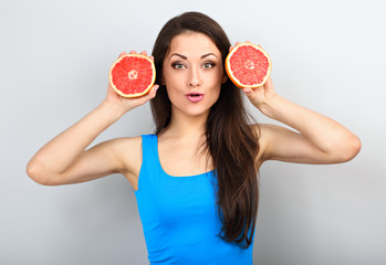 Beautiful funny grimacing brunette woman holding fresh red grapefruit near the face and looking humor on blue background.