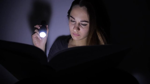 close-up portrait attentive young woman is reading a book with a flashlight in a dark room at night. 4k. Motion shooting